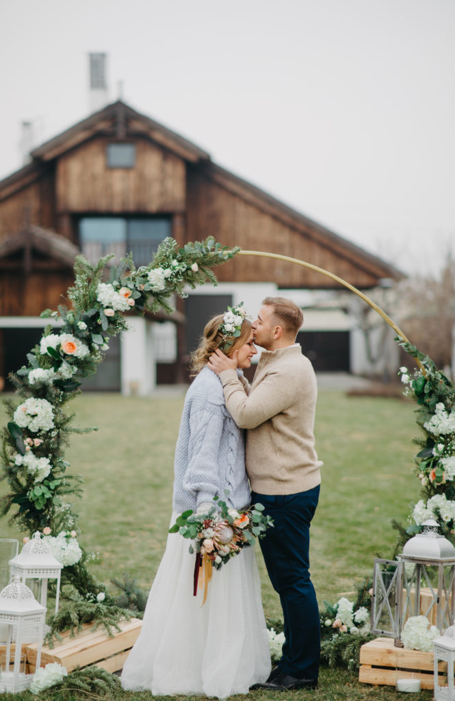 Couple sharing a forehead kiss in front of a circular arch decorated with flowers on their wedding day.